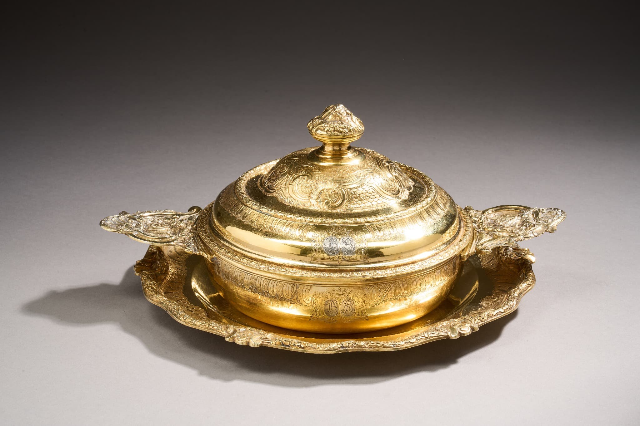 Ecuelle couverte et son plateau en vermeil. Le corps ciselé dans sa partie supérieure de canaux, feuillages stylisés, enroulements et rocailles. Les oreilles à contours sont à décor d’enroulements, coquilles déchiquetées, rocaille, feuilles d’acanthes et chutes de fleurs et feuillages. Le couvercle à double doucines présente deux ceintures de frises d’oves et de miroirs encadrant la même frise que le corps et au niveau de la terrasse une bande ciselée de rocaille et enroulements dans laquelle courent deux biches et un chien. La prise est constituée d’un bouton de feuillage et de coquilles. Le plateau circulaire et à contours présente une frise de canaux et de rocailles sur fond amati au niveau de l’aile. La bordure est ciselée d’une frise d’oves et de miroirs sur laquelle courent des branchages fleuris en alternance avec des agrafes à enroulement et fleurons. Les trois pièces sont gravées d’armoiries d’alliances (dégravées et regravées postérieurement d’un chiffre LB et GB sur un fond non vermeillé) sous une couronne de duc.