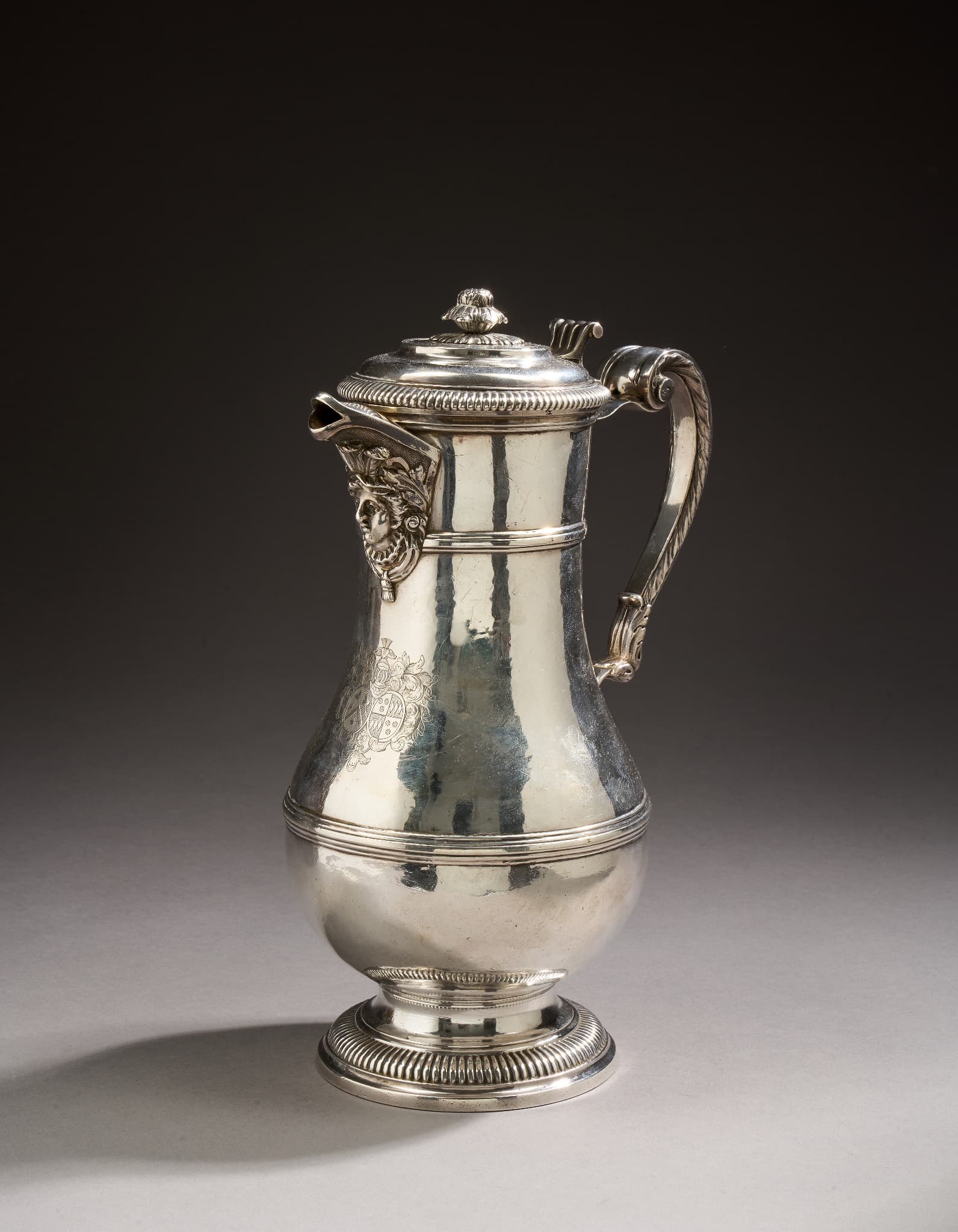 Aiguière en argent de forme balustre reposant sur une base moulurée de godrons? Le corps uni est ceinturé de deux joncs moulurés en applique. Le bec verseur est ciselé d’une tête d’espagnolette sur fond amati. Le couvercle à moulurations et doucines est bordé d’une frise de godrons, la prise est en graine éclatée. L’anse en console et ciselée de feuilles d’acanthes de refends. Le corps est gravé d’armoiries d’alliances sous un cimier (de face : marquis).