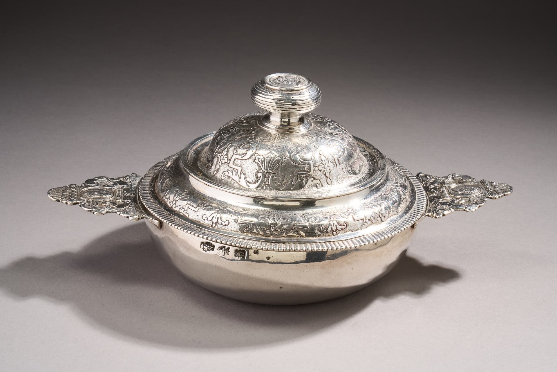 Ecuelle couverte en argent. Le corps uni, les oreilles à contours à décor de profil masculin et féminin à l’antique et de dauphins stylisés, coquilles et enroulements. Le couvercle bordé d’une frise de godrons et ciselé d’un double rang, d’enroulements, feuillages, palmettes et fleurons sur fond amati (petites usures). Le bouton orné de filets est surmonté d’un profil féminin à l’antique.