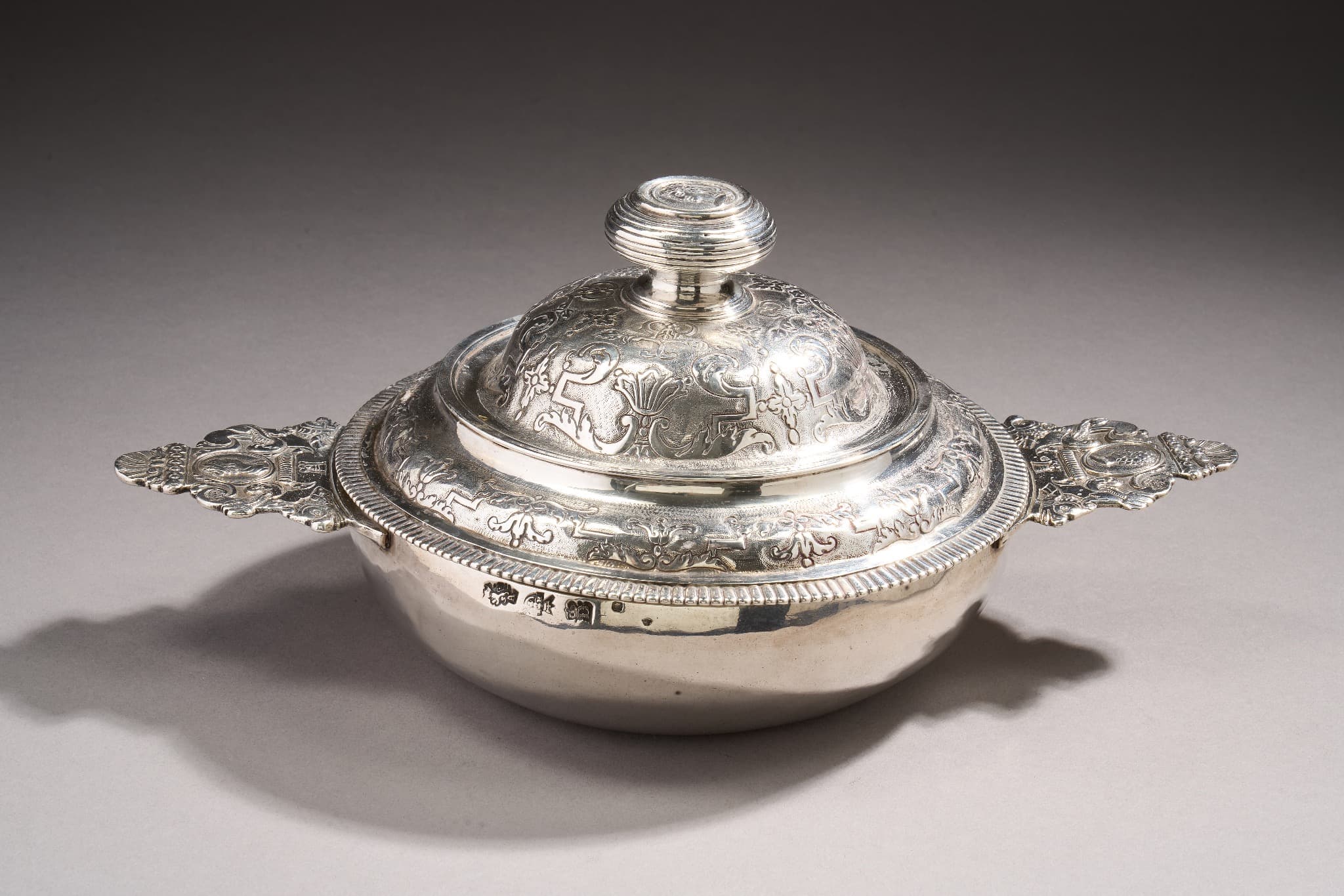 Ecuelle couverte en argent. Le corps uni, les oreilles à contours à décor de profil masculin et féminin à l’antique et de dauphins stylisés, coquilles et enroulements. Le couvercle bordé d’une frise de godrons et ciselé d’un double rang, d’enroulements, feuillages, palmettes et fleurons sur fond amati (petites usures). Le bouton orné de filets est surmonté d’un profil féminin à l’antique.