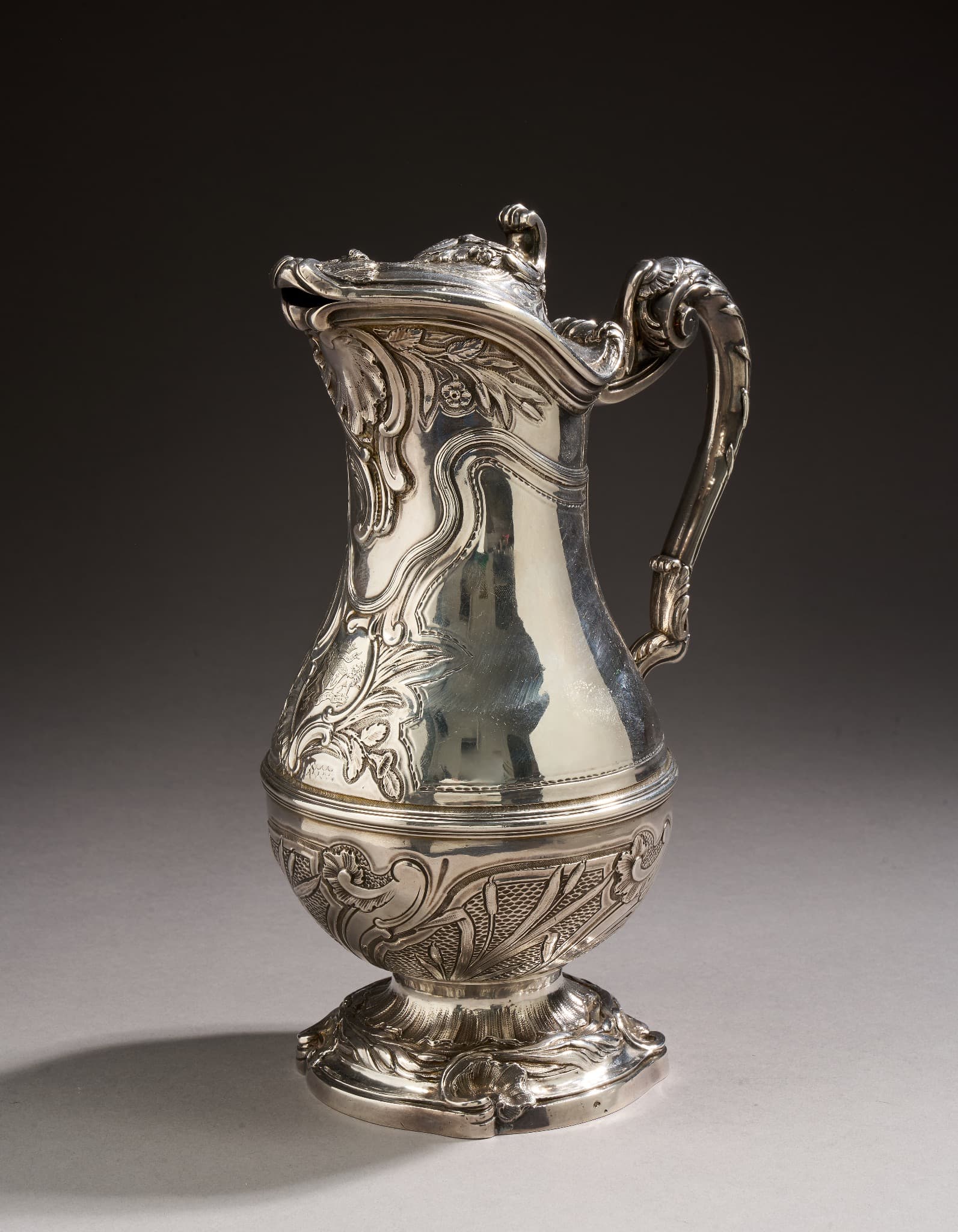 Aiguière en argent de forme balustre reposant sur un pied à contours moulurés et ciselés de coquilles déchiquetées, frises de roseaux et rocailles. La partie basse du corps est ciselée de compartiments, ornés de branchages de roseaux sur un fond ciselé imitant l’eau, et encadrés d’enroulements et de rocailles. Moulurations en partie médiane. La partie supérieure partiellement unie et ciselée d’un large motif d’enroulements, feuillages et fleurs. Motifs que nous retrouvons sous le bec verseur, ceux-là agrémentés de larges coquilles déchiquetées. Le couvercle à contours, ressauts et doucines présente les mêmes ornements. L’anse en console est ciselée d’enroulements, coquilles, feuilles d’acanthes et branchages de roseaux de refends. Le cartouche sur le corps gravé d’armoiries d’alliances soutenues par deux lions sous une couronne de marquis et de la devise “Victoria plus qu’un lion”