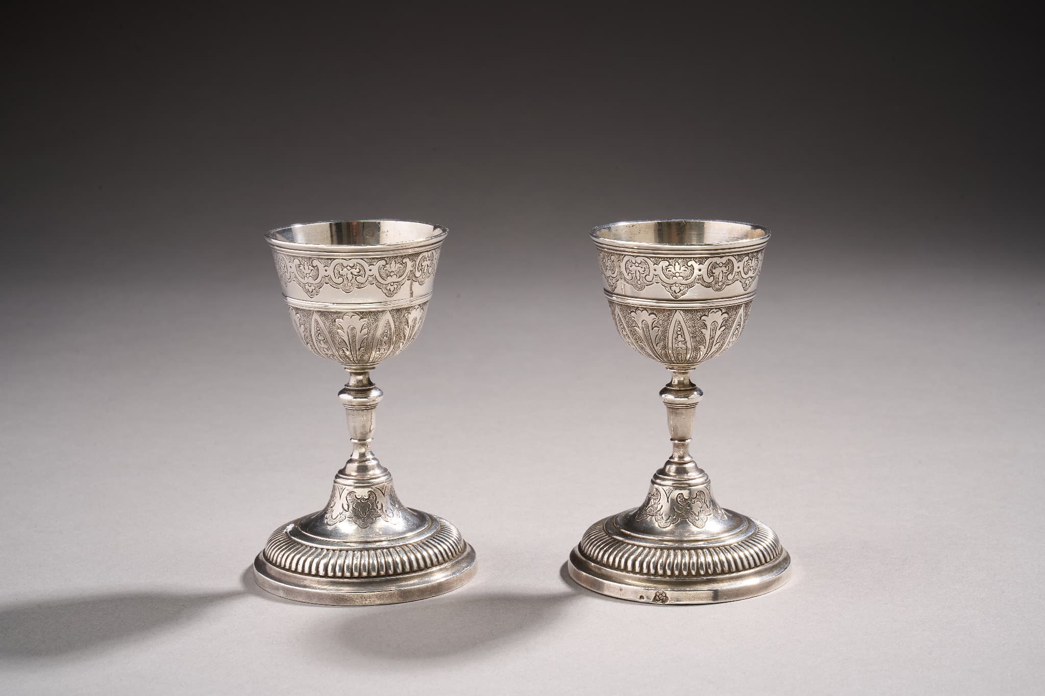 Paire de coquetiers en argent reposant sur une base circulaire à décor de godrons et d’une doucine ciselée de feuilles d’acanthes et fleurons sur fond amati. Le pied est légèrement renflé et la partie supérieure présente un décor en deux registres séparés par une bande de filets moulurés, d’une part de lambrequins et feuilles d’acanthes sur fond amati, d’autre part d’enroulements et de fleurons.
