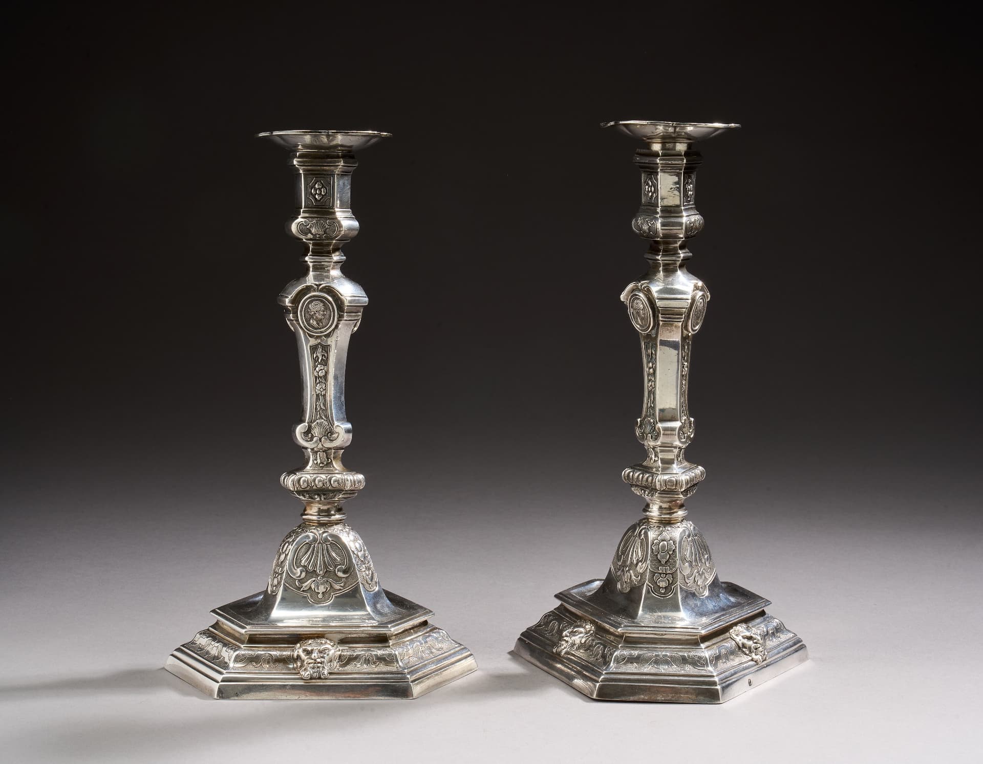Paire de flambeaux et leurs bobèches (non poinçonnées mais très probablement d’origine) en argent reposant sur une base triangulaire à ressauts et doucines ciselées d’une frise d’enroulements, fleurons et croisillons sur fond amati. La cloche est ciselée de coquilles, fleurettes, fleurons et écailles sur font amati. Le fût, triangulaire, ciselé de frises d’oves, chutes de fleurons, coquilles et profils à l’antique. Le binet à décor de compartiments, enroulements, coquilles et fleurons sur fond amati. Les bobèches polylobées.