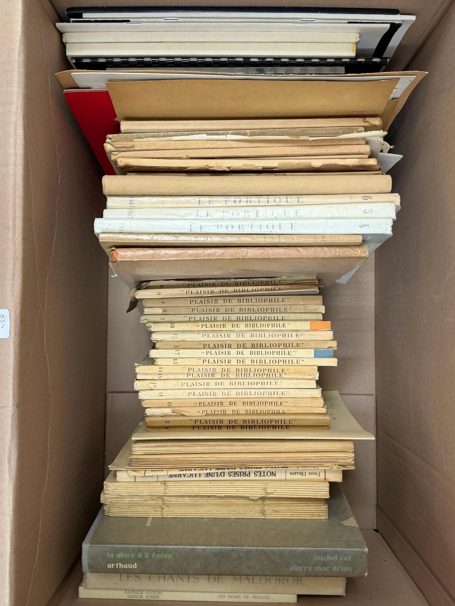 Littérature. Ensemble de livres et brochures, principalement brochés, de formats variés, rangés dans un carton. Titres visibles : 'NOTES PRISES D'UNE LÉGION', 'PLAISIR DE BIBLIOPHILE', 'LE FONIOLET', 'LES GÉANTS DE BALADOUC', sans autres informations bibliographiques certaines. Nombreux exemplaires en état d’usage, certains avec dos abîmés ou froissés.