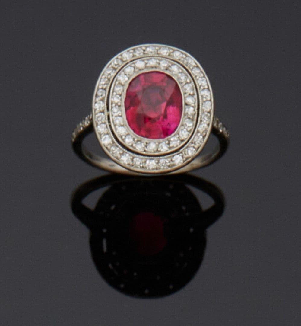 Bague en platine (850e), centrée d'un rubis ovale facetté en serti clos perlé, dans un double entourage de diamants taille 8/8. Le panier ajouré décoré de volutes. Vers 1920. Intérieur numéroté
