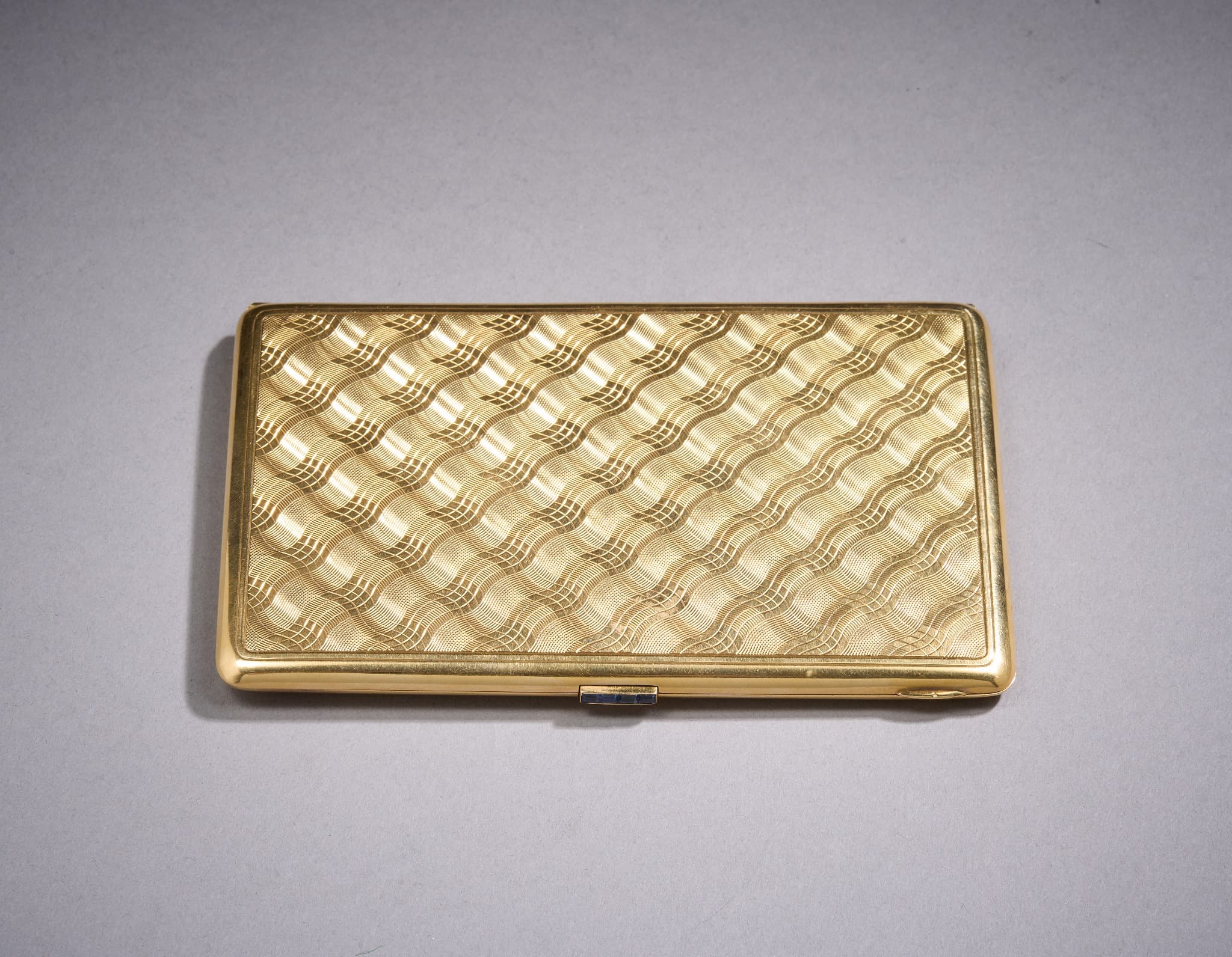 Étui à cigarettes en or (750°/°°) à décor guilloché de vagues s’entrecroisant. Le bouton d'ouverture serti de calibrés de saphirs. La barrette interne à décor repercé de fleurettes.